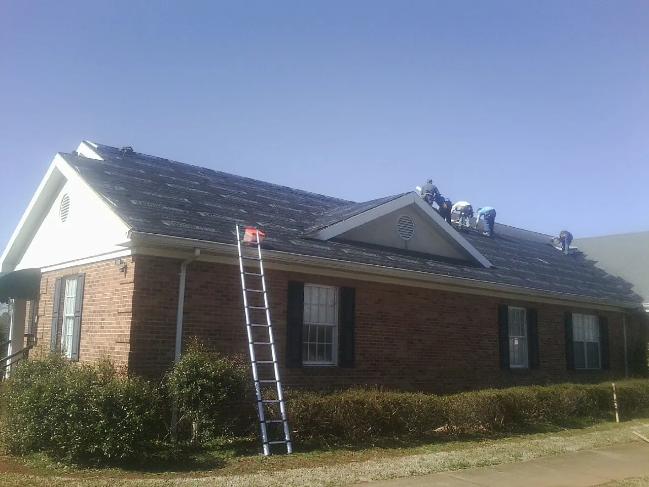 Crew tearing shingles off a brick ranch roof with a ladder set — Metal Roof Repair work by our crew in Manson