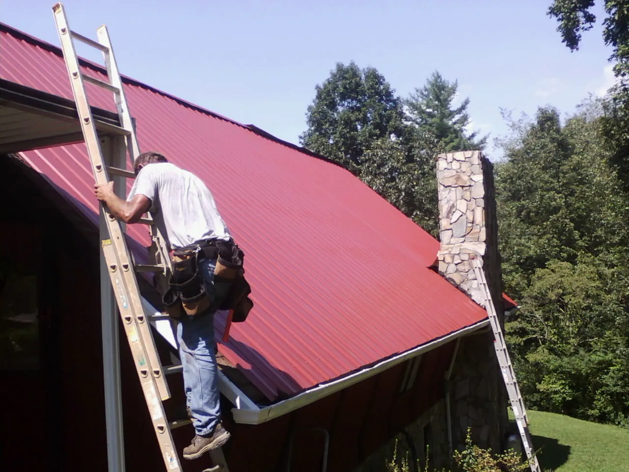 Roofer on a ladder beside a red metal roof and stone chimney — Soffit & Fascia Repair by Roofing Brothers in Manson